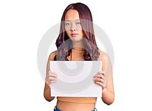 Young beautiful chinese girl holding paper banner with blank space thinking attitude and sober expression looking self confident