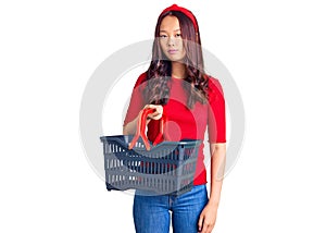 Young beautiful chinese girl holding supermarket shopping basket thinking attitude and sober expression looking self confident