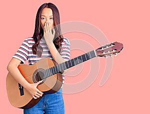 Young beautiful chinese girl playing classical guitar covering mouth with hand, shocked and afraid for mistake