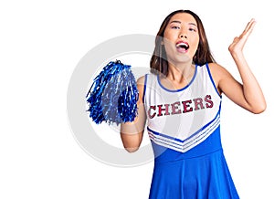 Young beautiful chinese girl wearing cheerleader uniform holding pompom celebrating victory with happy smile and winner expression