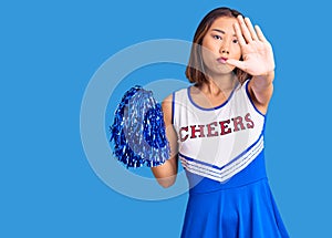 Young beautiful chinese girl wearing cheerleader uniform holding pompom with open hand doing stop sign with serious and confident