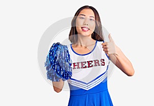 Young beautiful chinese girl wearing cheerleader uniform holding pompom smiling happy and positive, thumb up doing excellent and