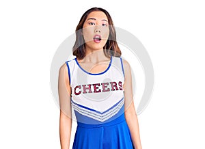 Young beautiful chinese girl wearing cheerleader uniform in shock face, looking skeptical and sarcastic, surprised with open mouth