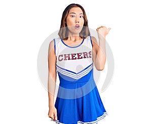Young beautiful chinese girl wearing cheerleader uniform surprised pointing with hand finger to the side, open mouth amazed