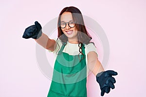 Young beautiful chinese girl wearing cleaner apron looking at the camera smiling with open arms for hug