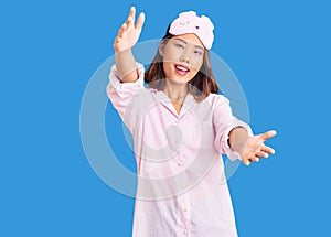 Young beautiful chinese girl wearing sleep mask and pajama looking at the camera smiling with open arms for hug