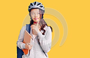 Young beautiful chinese girl wearing student backpack and bike helmet holding book serious face thinking about question with hand