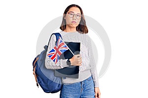 Young beautiful chinese girl wearing student backpack holding binder and uk flag thinking attitude and sober expression looking