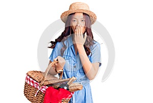 Young beautiful chinese girl wearing summer hat and holding picnic wicker basket with bread covering mouth with hand, shocked and