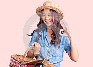 Young beautiful chinese girl wearing summer hat and holding picnic wicker basket with bread doing ok sign with fingers, smiling