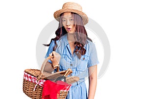 Young beautiful chinese girl wearing summer hat and holding picnic wicker basket with bread scared and amazed with open mouth for
