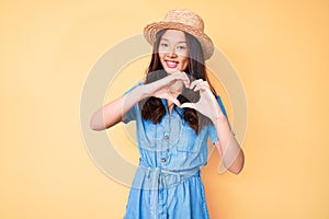 Young beautiful chinese girl wearing summer hat smiling in love doing heart symbol shape with hands