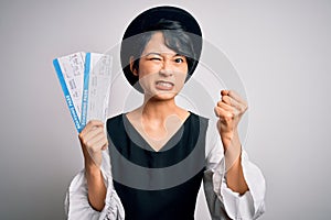 Young beautiful chinese tourist woman holding boarding pass airlane over white background annoyed and frustrated shouting with