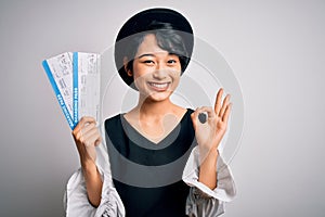 Young beautiful chinese tourist woman holding boarding pass airlane over white background doing ok sign with fingers, excellent