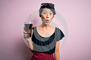 Young beautiful chinese woman drinking cola fizzy beverage using straw over pink background scared in shock with a surprise face,