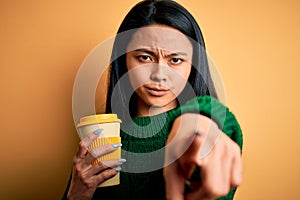 Young beautiful chinese woman drinking cup of coffee over isolated yellow background pointing with finger to the camera and to
