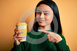 Young beautiful chinese woman drinking cup of coffee over isolated yellow background very happy pointing with hand and finger