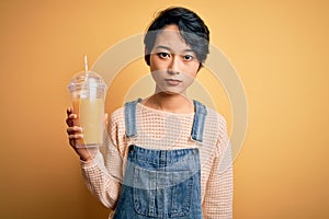 Young beautiful chinese woman drinking healthy orange juice over isolated yellow background with a confident expression on smart