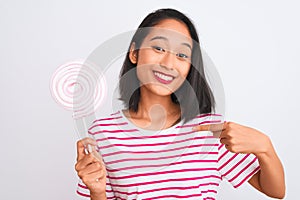 Young beautiful chinese woman eating lollipop standing over isolated white background with surprise face pointing finger to