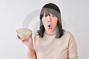 Young beautiful Chinese woman holding bowl with rice over isolated white background scared in shock with a surprise face, afraid