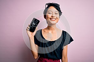 Young beautiful chinese woman holding broken smartphone showing craked screen with a happy face standing and smiling with a