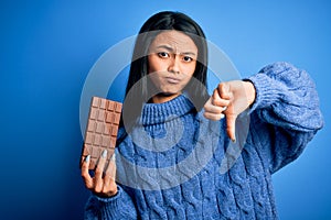 Young beautiful chinese woman holding chocolate bar over isolated blue background with angry face, negative sign showing dislike