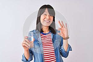 Young beautiful Chinese woman holding glass of water over isolated white background doing ok sign with fingers, excellent symbol