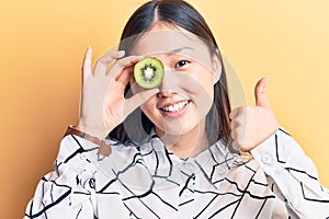 Young beautiful chinese woman holding kiwi over eye smiling happy and positive, thumb up doing excellent and approval sign