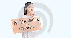 Young beautiful chinese woman holding mother nature is crying cardboard banner serious face thinking about question with hand on