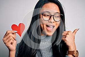 Young beautiful chinese woman holding paper heart over isolated white background pointing and showing with thumb up to the side