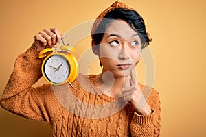 Young beautiful chinese woman holding vintage alarm clock over isolated yellow background serious face thinking about question,