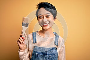 Young beautiful chinese woman painting holding paint brush over isolated yellow background with a happy face standing and smiling