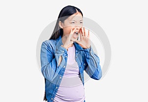 Young beautiful chinese woman pregnant expecting baby shouting angry out loud with hands over mouth