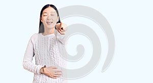 Young beautiful chinese woman wearing casual sweater laughing at you, pointing finger to the camera with hand over body, shame