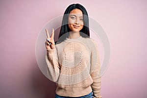 Young beautiful chinese woman wearing casual sweater over isolated pink background showing and pointing up with fingers number two