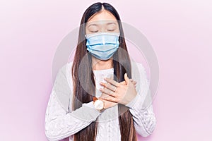 Young beautiful chinese woman wearing medical mask smiling with hands on chest, eyes closed with grateful gesture on face
