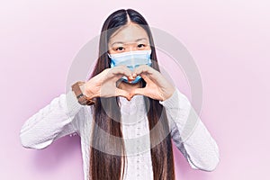 Young beautiful chinese woman wearing medical mask smiling in love doing heart symbol shape with hands