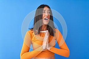 Young beautiful chinese woman wearing orange t-shirt standing over isolated blue background begging and praying with hands