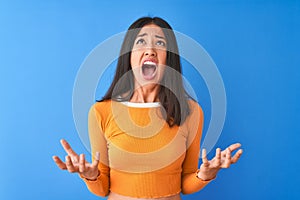 Young beautiful chinese woman wearing orange t-shirt standing over isolated blue background crazy and mad shouting and yelling