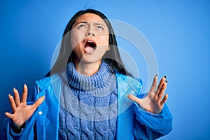 Young beautiful chinese woman wearing rain coat standing over isolated blue background crazy and mad shouting and yelling with