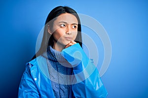 Young beautiful chinese woman wearing rain coat standing over isolated blue background with hand on chin thinking about question,