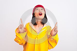 Young beautiful chinese woman wearing raincoat and wool cap over isolated white background amazed and surprised looking up and