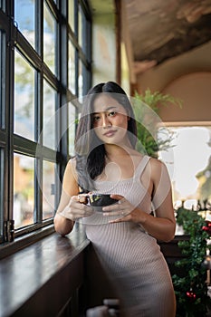 Young beautiful Asian woman having a coffee break while leaning against the windows