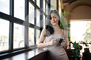 Young beautiful Asian woman having a coffee break while leaning against the windows