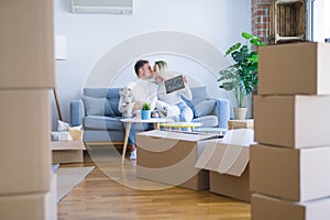 Young beautiful couple with dog sitting on the sofa holding blackboard with message kissing at new home around cardboard boxes