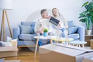 Young beautiful couple with dog sitting on the sofa holding blackboard with message at new home around cardboard boxes