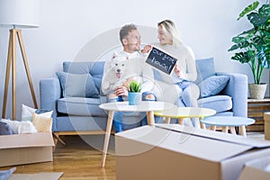 Young beautiful couple with dog sitting on the sofa holding blackboard with message at new home around cardboard boxes