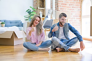 Young beautiful couple moving to a new house sitting on the floor doing happy thumbs up gesture with hand