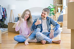 Young beautiful couple moving to a new house sitting on the floor doing happy thumbs up gesture with hand