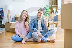 Young beautiful couple moving to a new house sitting on the floor smiling with happy face looking and pointing to the side with